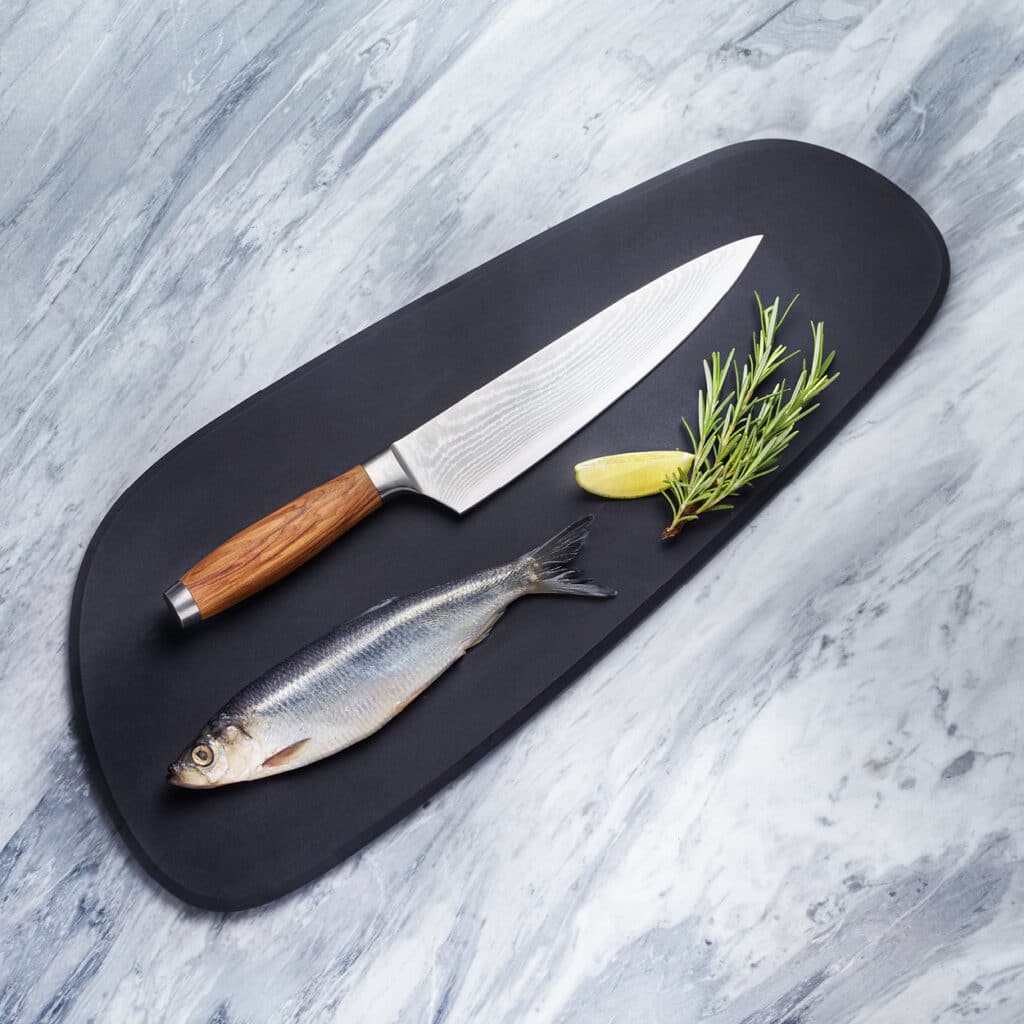 Santokumesser mit Holzgriff auf schwarzem Board neben frischem Fisch, Rosmarinzweig und Zitronenspalte.