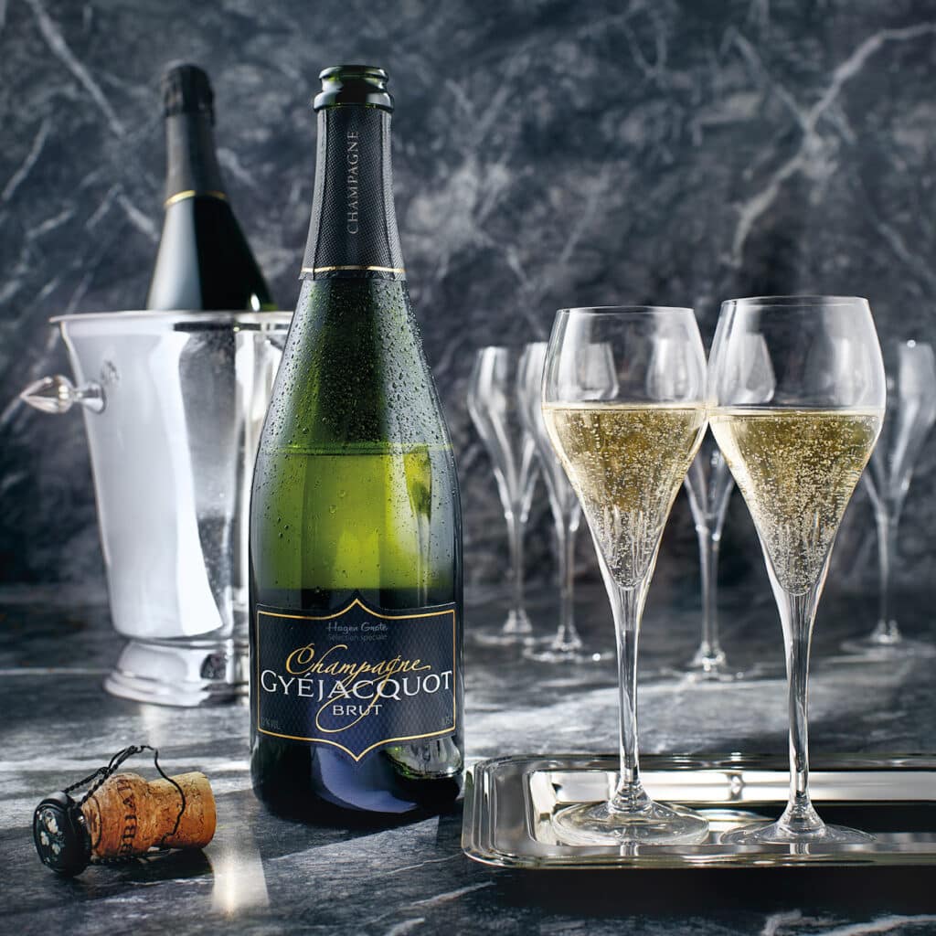 Champagnerflasche und zwei gefüllte Sektgläser auf Marmortisch mit Kühler im Hintergrund.