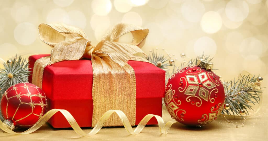 Rotes Weihnachtsgeschenk mit goldener Schleife und glitzernden Christbaumkugeln vor goldenem Bokeh-Hintergrund.